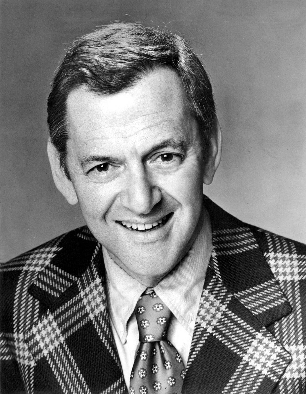 Peter Finch as Howard Beale, illuminated by TV studio lights, with his mouth open in a frantic yell during a broadcast.